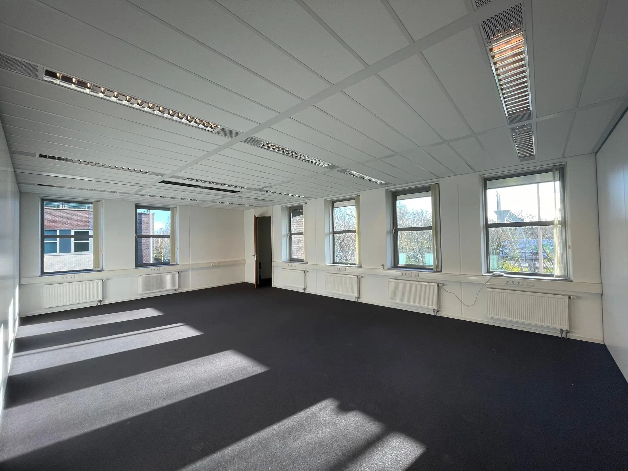 Lichte, lege kantoorruimte met vijf ramen, verlaagd plafond en donkere vloerbedekking.